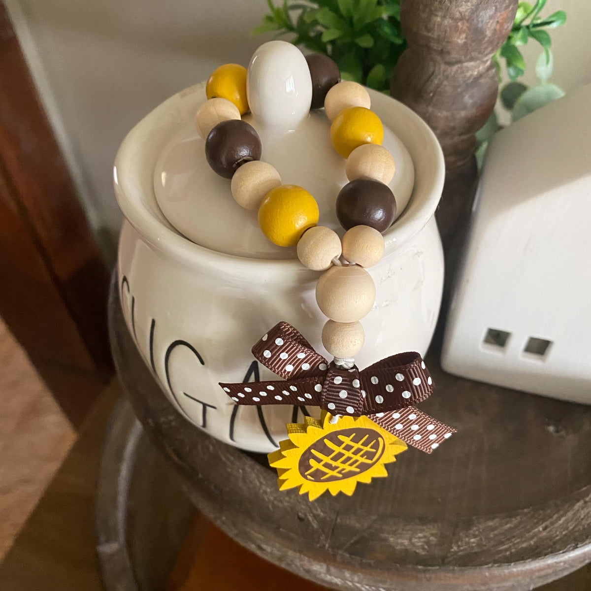 A decorative beaded garland with natural wood, mustard yellow, and dark brown beads draped over a white ceramic canister, featuring a brown polka-dot ribbon bow and a yellow sunflower charm.