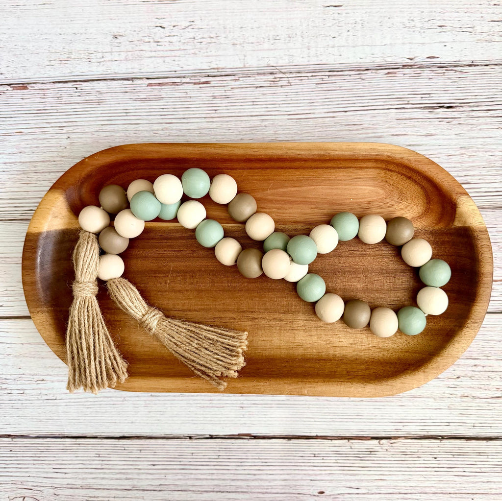 A wooden bead garland with two tassels, featuring matte beads in sage green, cream, and light brown, displayed on a wooden serving tray over a white-washed wood background.