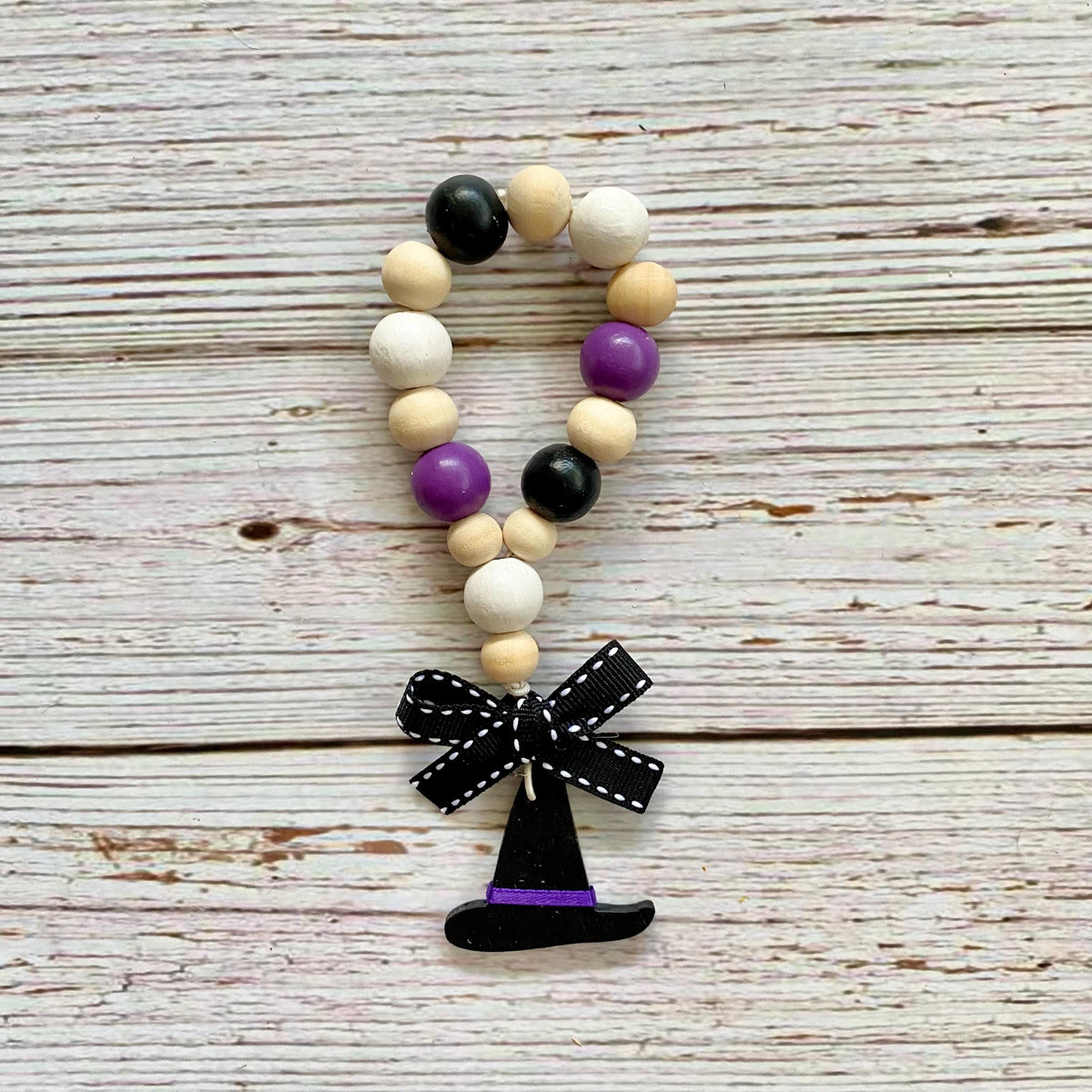 A beaded garland with alternating black, cream, and purple wooden beads, featuring a black ribbon bow with white polka dots and a small witch hat charm, displayed on a rustic whitewashed wood background.