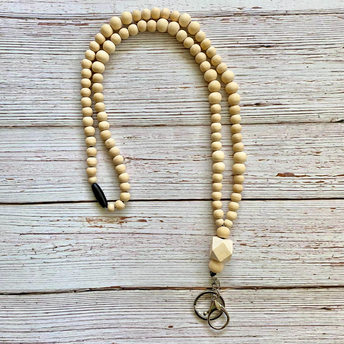 A long strand of light natural wood bead lanyard, laid out on a whitewashed wooden surface. The strand features a small black breakaway clasp near the top left and a faceted light wood accent bead near the bottom right, ending in a silver-toned metal clasp with two rings.
