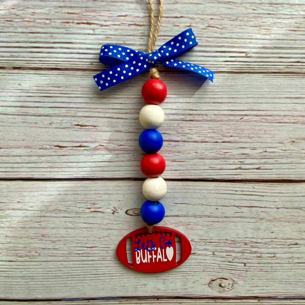 A patriotic beaded ornament with red, white, and blue wooden beads, a blue polka-dot ribbon bow, and a red oval tag that reads 'SOUTHEASTERN BUFFALO' on a rustic white wood plank background.