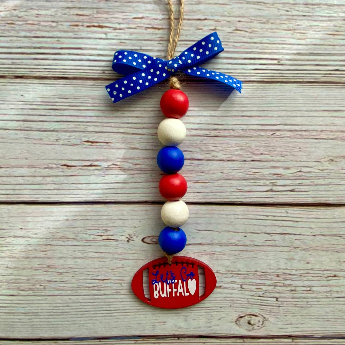 A patriotic beaded ornament with red, white, and blue wooden beads, a blue polka-dot ribbon bow, and a red oval tag that reads 'SOUTHEASTERN BUFFALO' on a rustic white wood plank background.