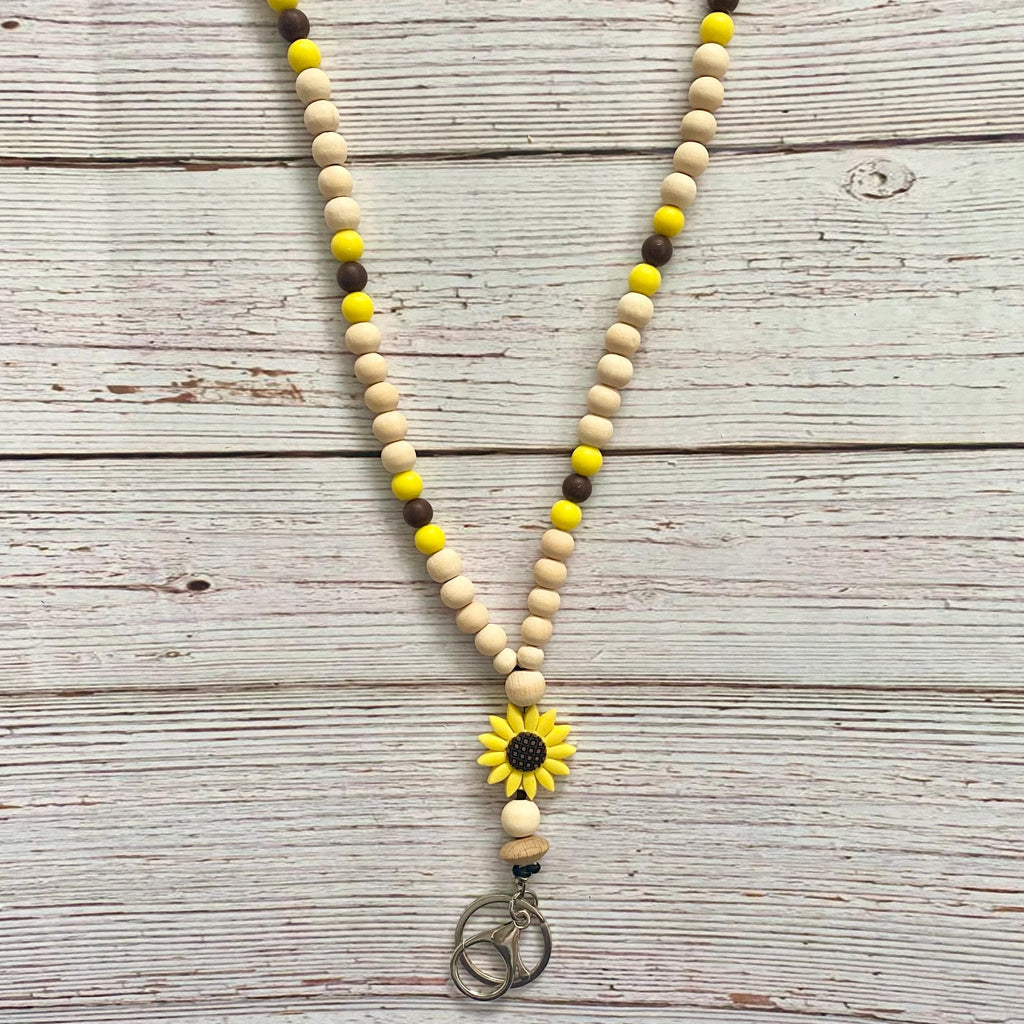 A wooden bead necklace with natural beige beads, yellow accent beads, and dark brown accent beads, featuring a yellow sunflower charm and a silver-toned peace sign pendant, laid flat on a whitewashed wooden background.