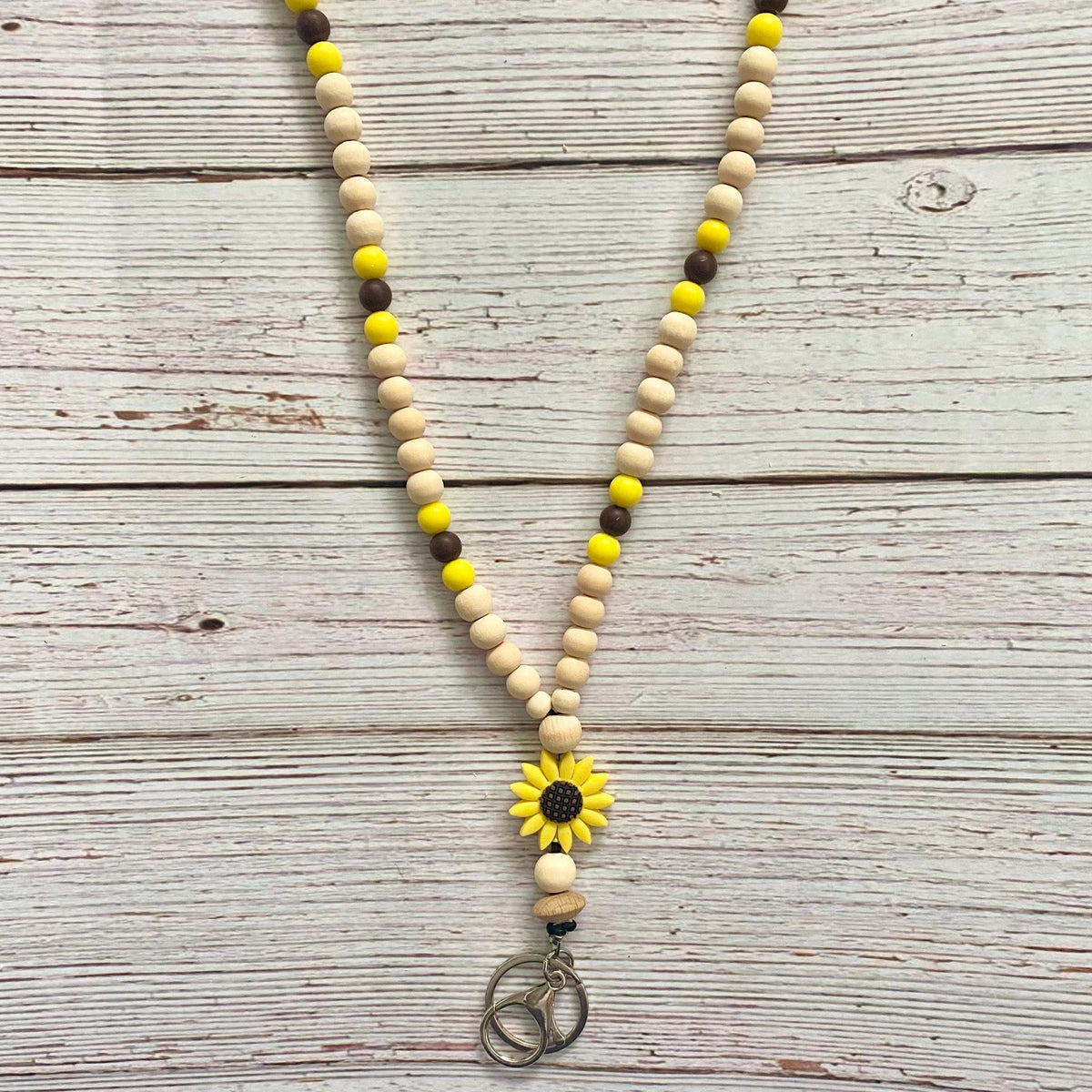A wooden bead necklace with natural beige beads, yellow accent beads, and dark brown accent beads, featuring a yellow sunflower charm and a silver-toned peace sign pendant, laid flat on a whitewashed wooden background.