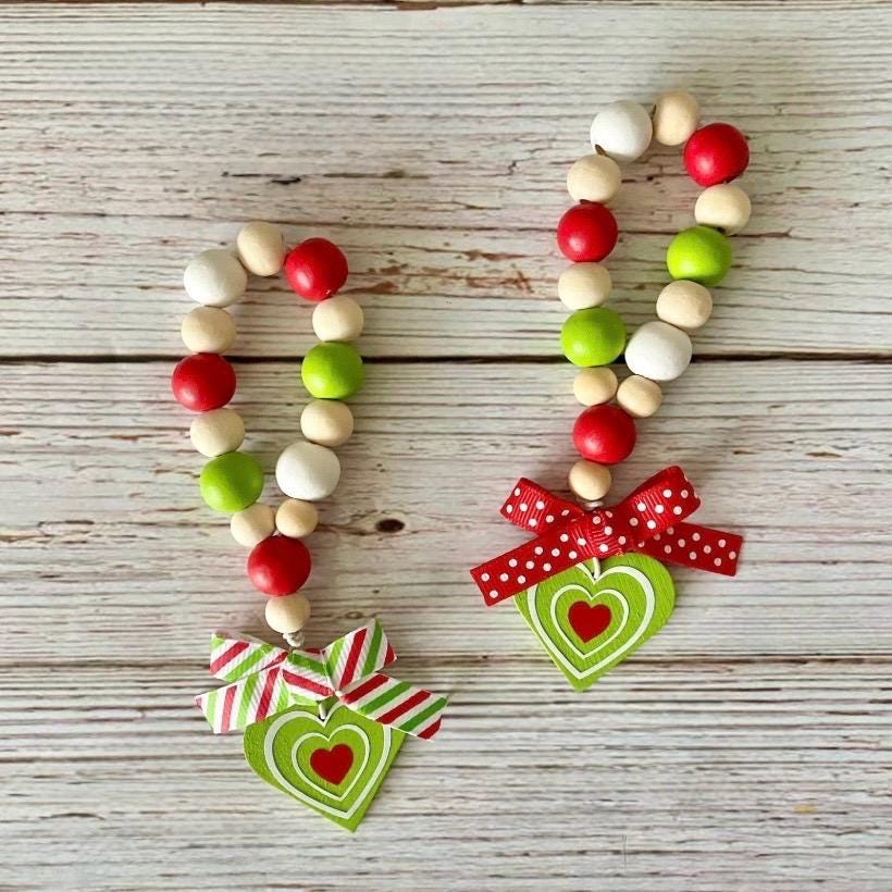 Two wooden bead garlands with red and green beads, decorated with red and white polka dot bows and green heart ornaments with red hearts, displayed on a rustic white-washed wooden background.