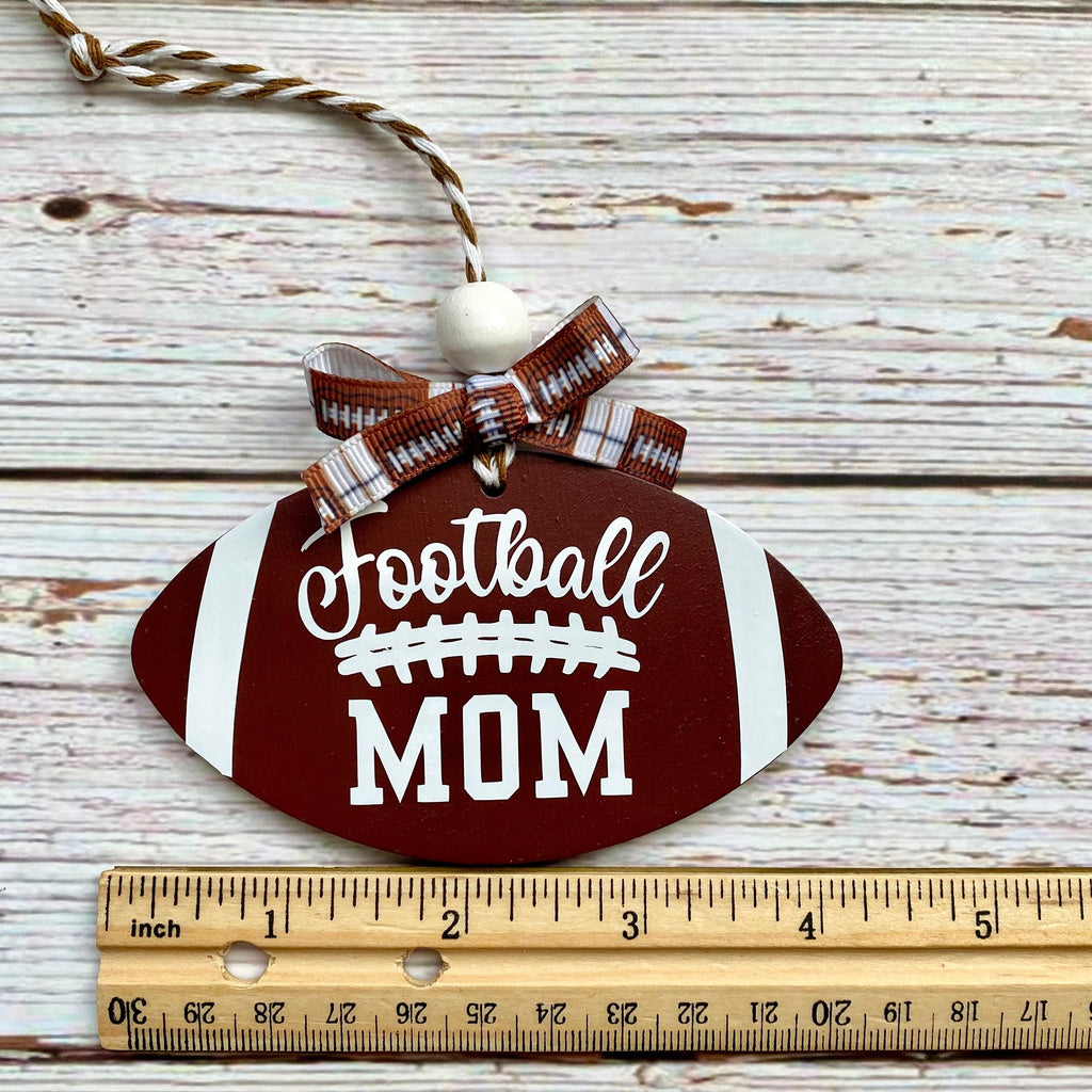 A football-shaped wooden ornament with 'Football MOM' printed on it, hanging on a whitewashed wood surface with a wooden ruler below for scale.