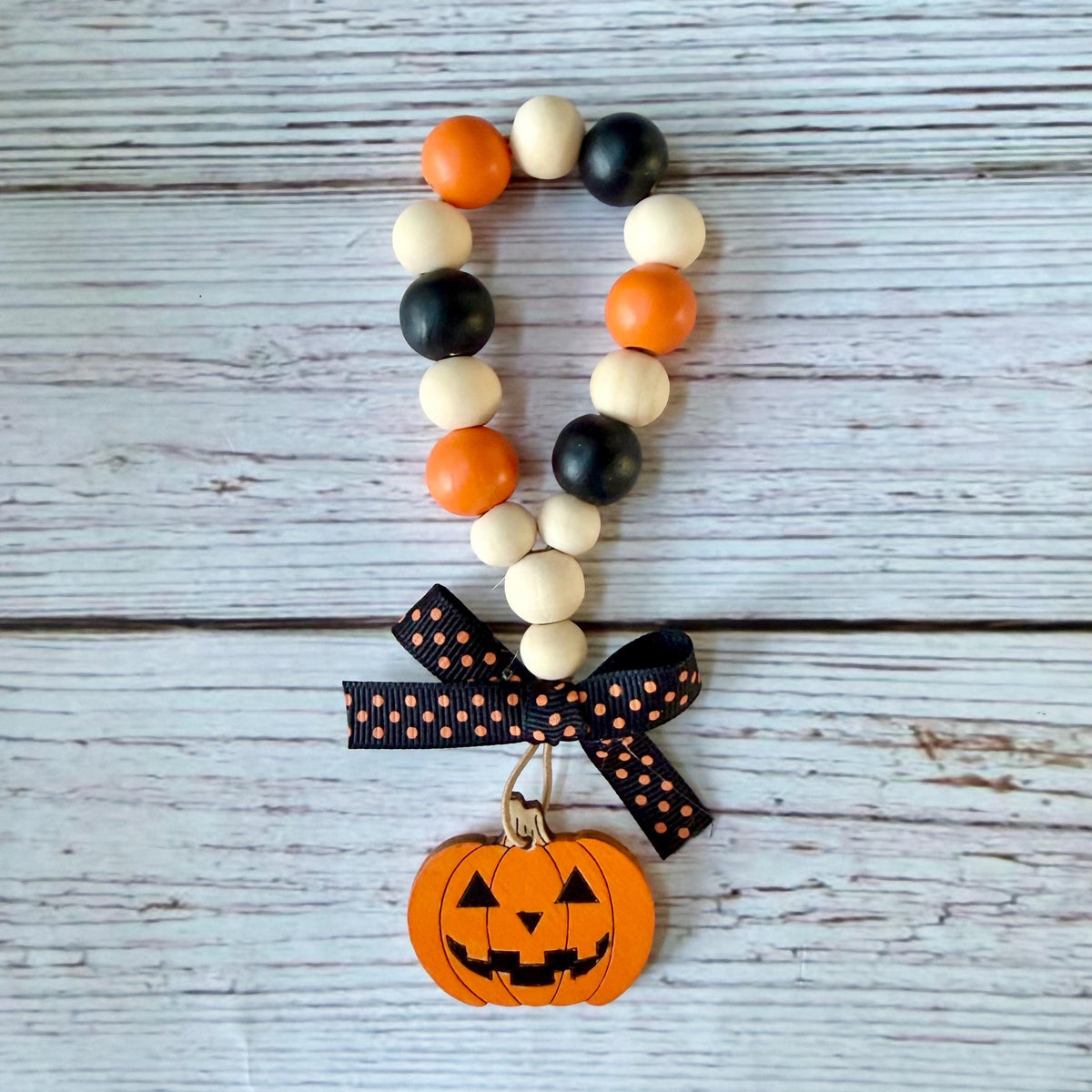 A wooden bead garland with orange, black, and cream beads, finished with a black ribbon printed with orange hearts and a hanging orange jack-o'-lantern charm, displayed on a rustic white-washed wood background.