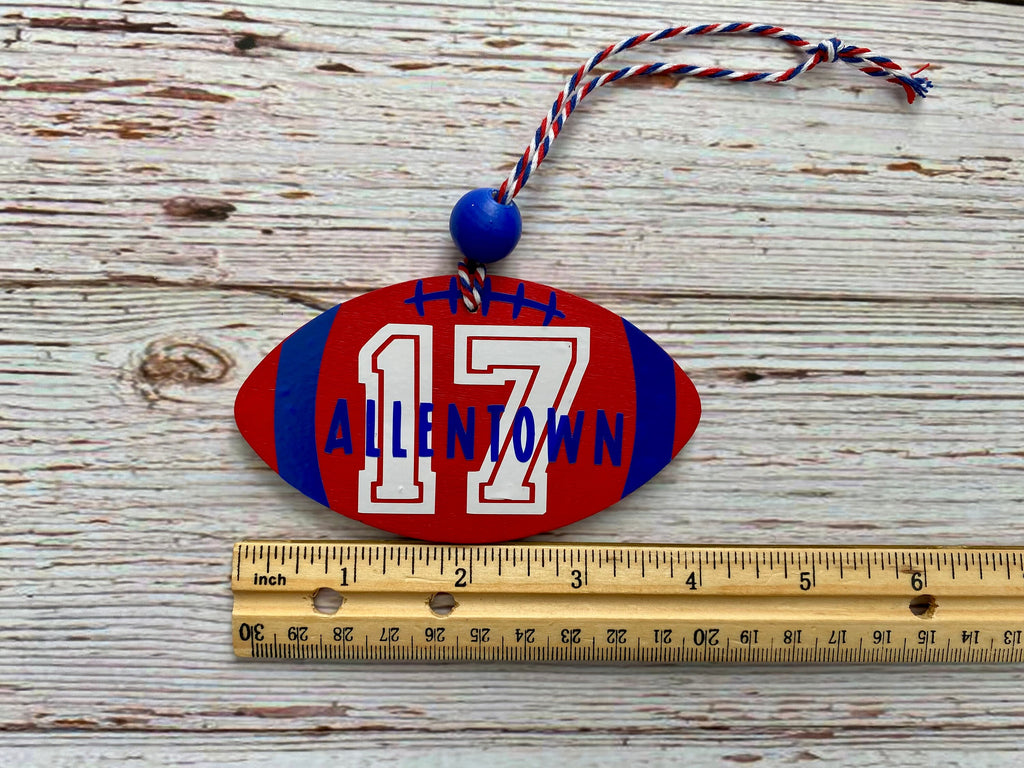 A football-shaped personalized ornament with the name 'ALLENTOWN' and number '17' in red, white, and blue, shown on a rustic wood background with a wooden ruler for scale.