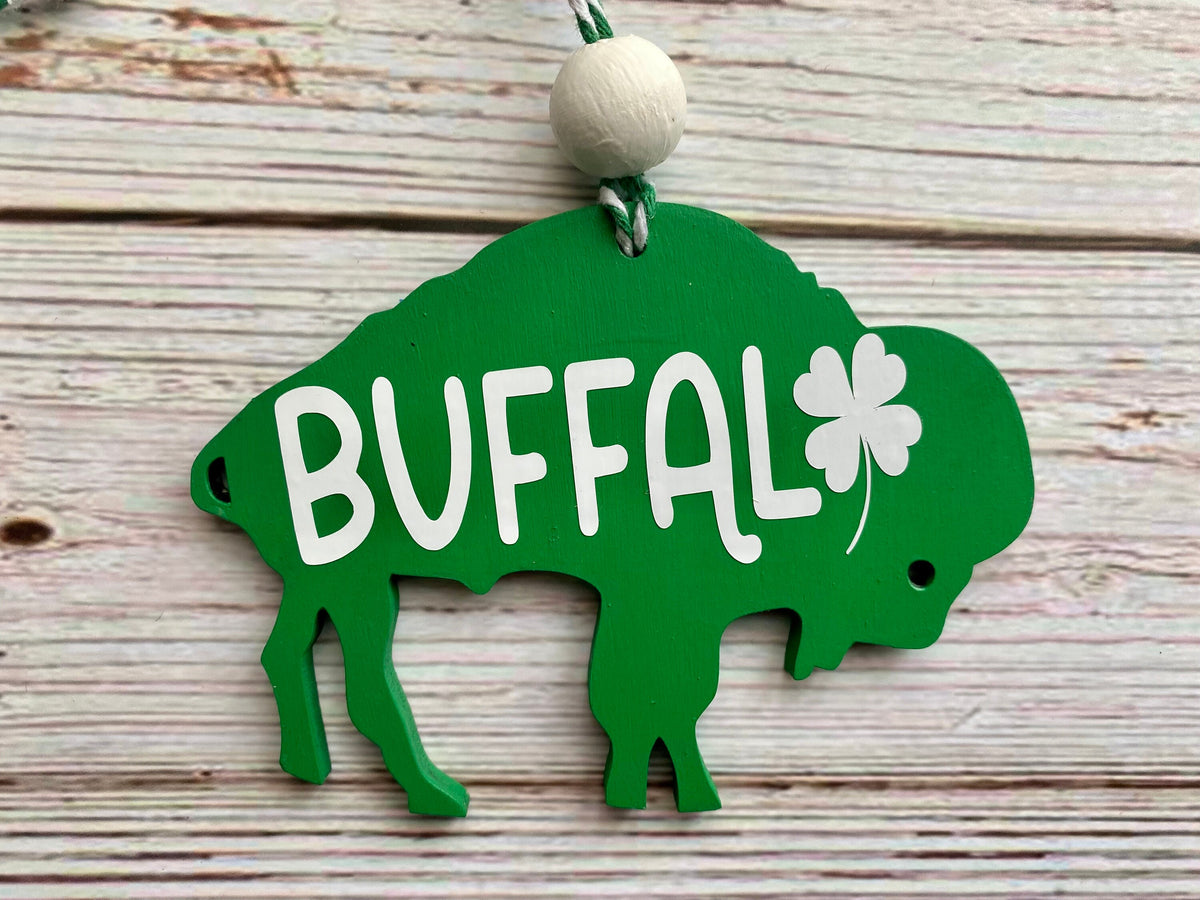 A green wooden St. Patrick's Day buffalo ornament with the word 'BUFFAL' and a shamrock cut out, hanging from a white bead on a whitewashed wood background.