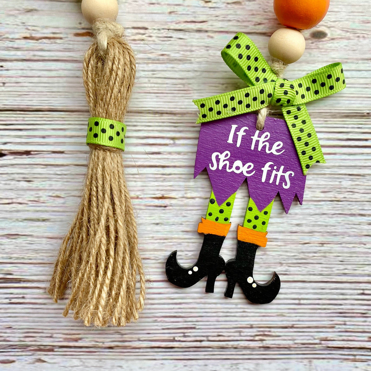 Two Halloween-themed wooden bead garlands on a whitewashed wood background. One ornament features a jute tassel with a green polka dot band, and the other features a purple sign reading 'If the shoe fits' with a green polka dot bow, orange accents, and black witch boots.