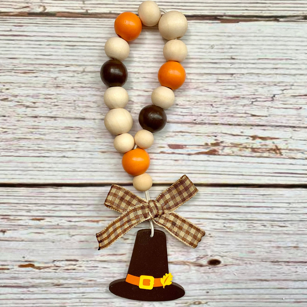 A fall-themed wooden bead garland with natural wood, orange, and dark brown beads, finished with a brown plaid ribbon bow and a brown pilgrim hat charm with a yellow belt.