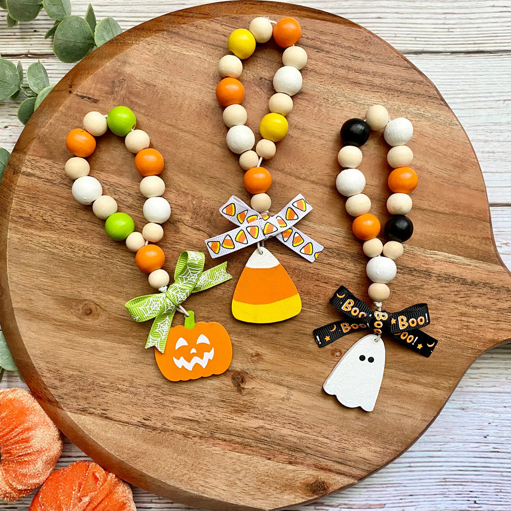 Three beaded Halloween-themed bead loops displayed on a round wooden serving platter. The keychains feature natural wood beads with orange, white, yellow, green, and black accents, decorated with autumn charms including a jack-o'-lantern, candy corn, a windmill, and a ghost.
