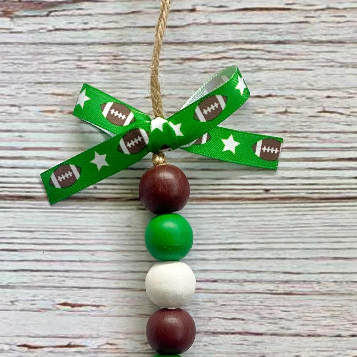 A decorative hanging ornament with a green ribbon bow printed with footballs and stars, suspended from a natural twine string. Below the bow are four round beads in brown, green, white, and brown, arranged vertically on a whitewashed wood background.