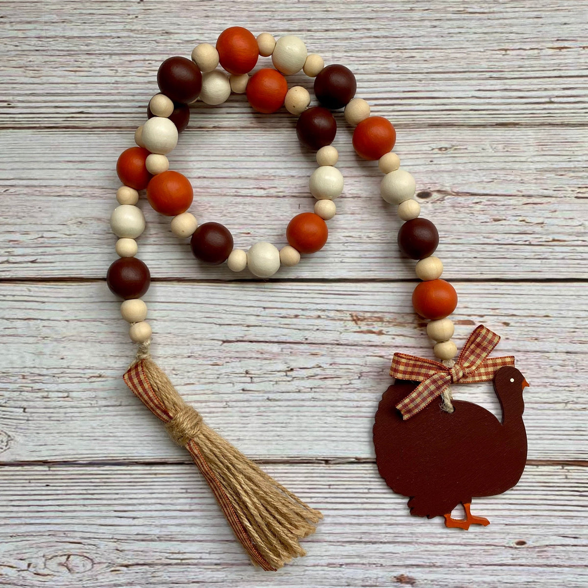 A rustic autumn-themed bead garland with orange, brown, and cream wooden beads, a jute tassel, and a dark brown turkey silhouette ornament with a gingham bow, displayed on a whitewashed wood plank background.