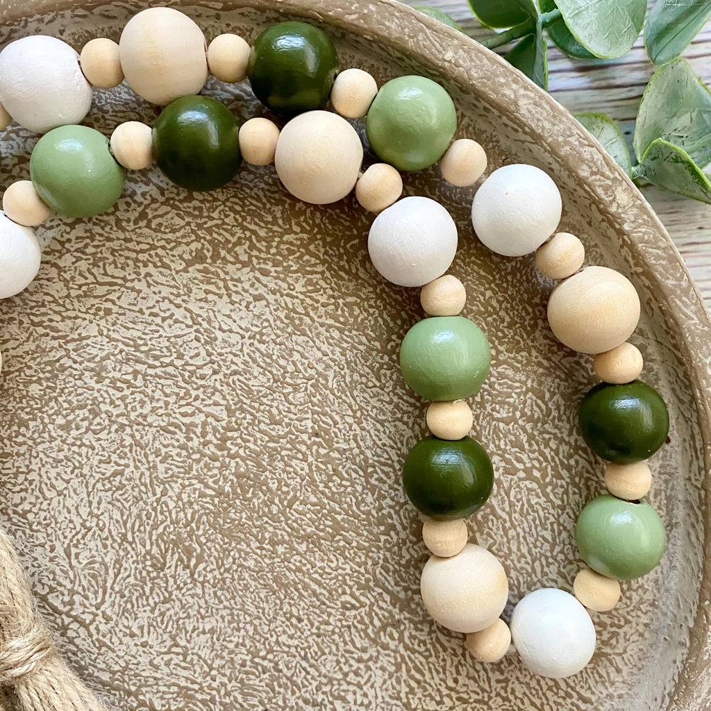 A beaded garland with large wooden beads in natural cream, light sage green, and dark olive green, arranged on a textured beige ceramic plate.