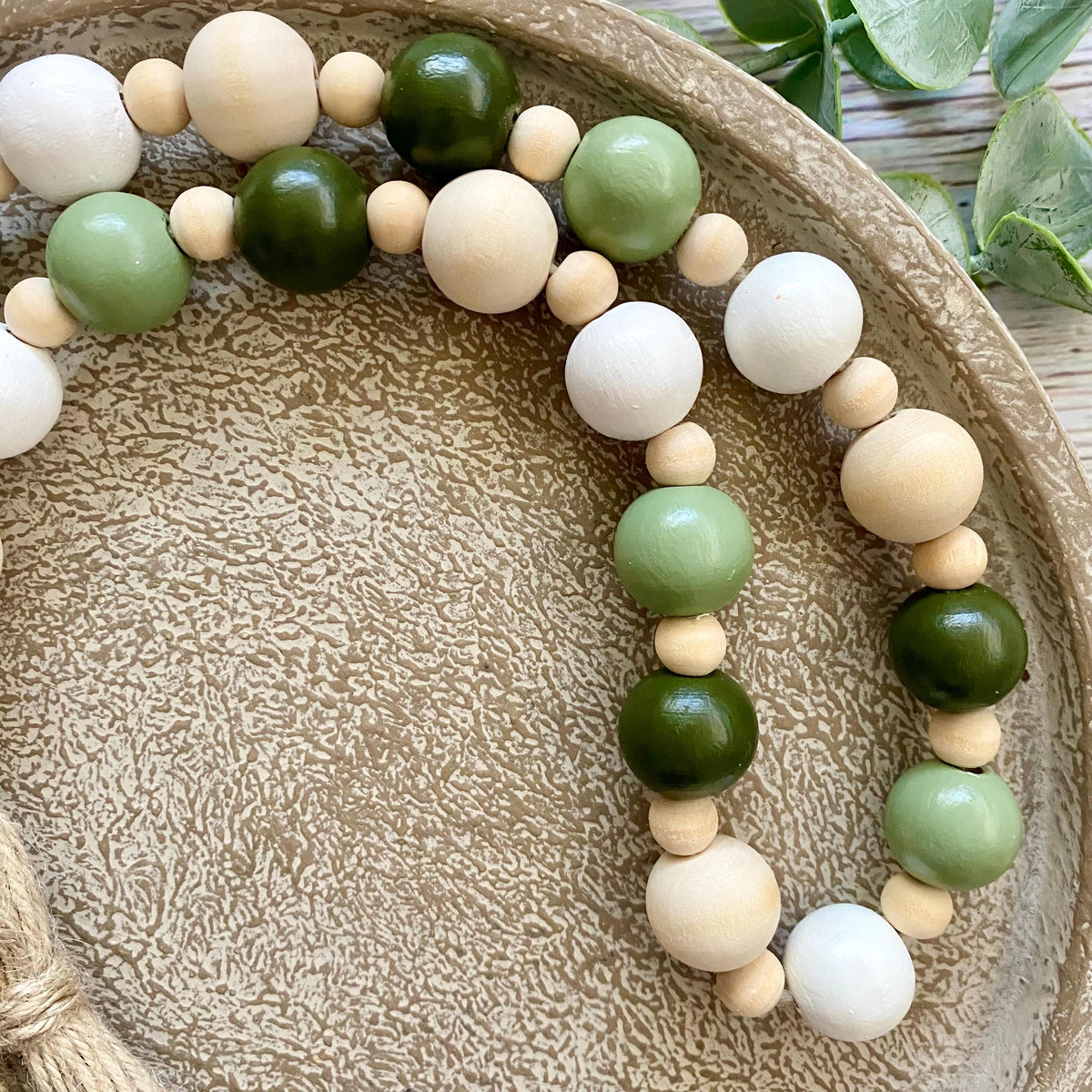 A beaded garland with large wooden beads in natural cream, light sage green, and dark olive green, arranged on a textured beige ceramic plate.