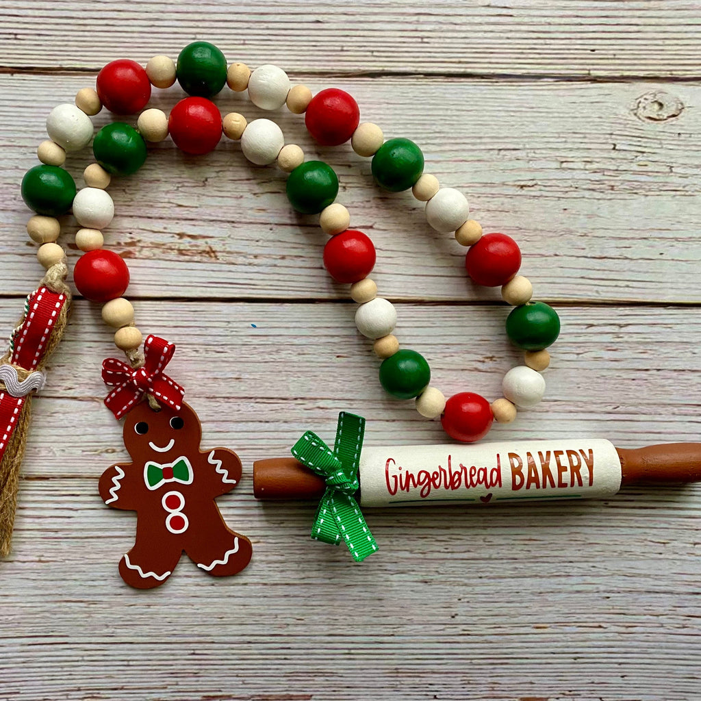 A Christmas-themed wooden bead garland with red, green, and white beads, a gingerbread man ornament, a tassel, and a gingerbread rolling pin that reads 'Gingerbread BAKERY', displayed on a light wood background.