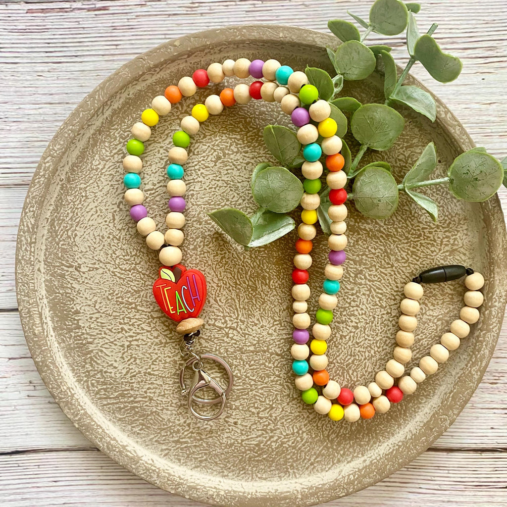 A colorful wooden bead necklace with a red heart charm and silver keyring, displayed on a textured beige tray with green eucalyptus leaves.