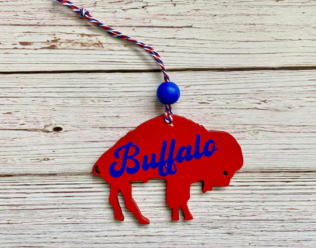 A red buffalo silhouette ornament with the word 'Buffalo' in blue script, hanging from a red, white, and blue braided cord with a blue bead, displayed on a white wood plank background.