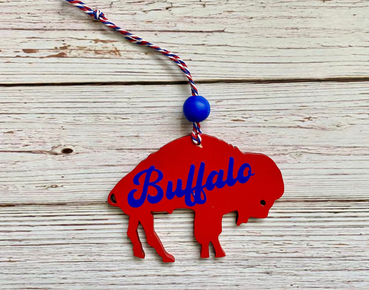 A red buffalo silhouette ornament with the word 'Buffalo' in blue script, hanging from a red, white, and blue braided cord with a blue bead, displayed on a white wood plank background.