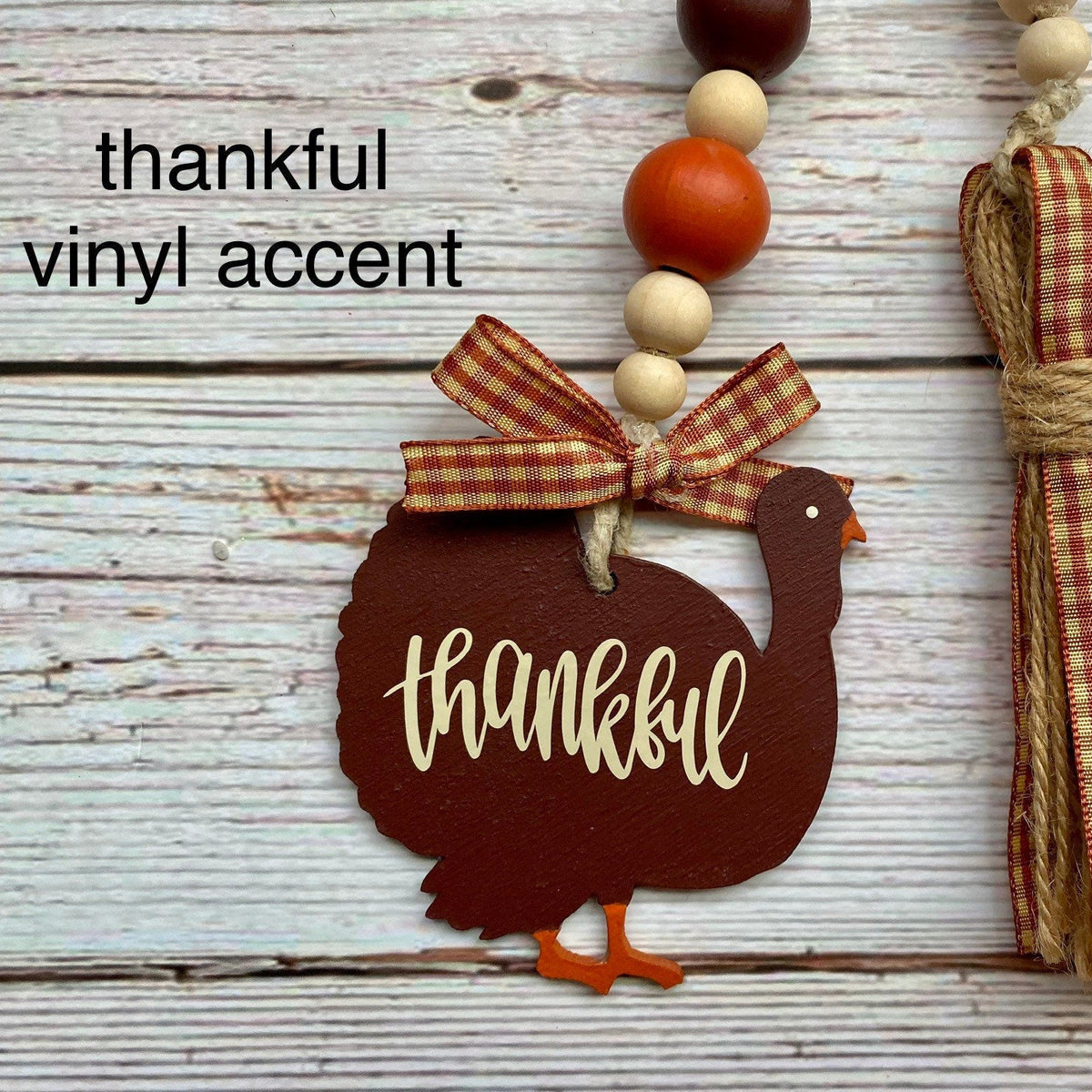 A rustic decorative hanging ornament with a turkey-shaped wooden tag reading “thankful” in white script, attached to a beaded garland with a red-and-beige gingham ribbon bow. A matching tassel with the same gingham ribbon and tan cord hangs beside it on a whitewashed wood surface.