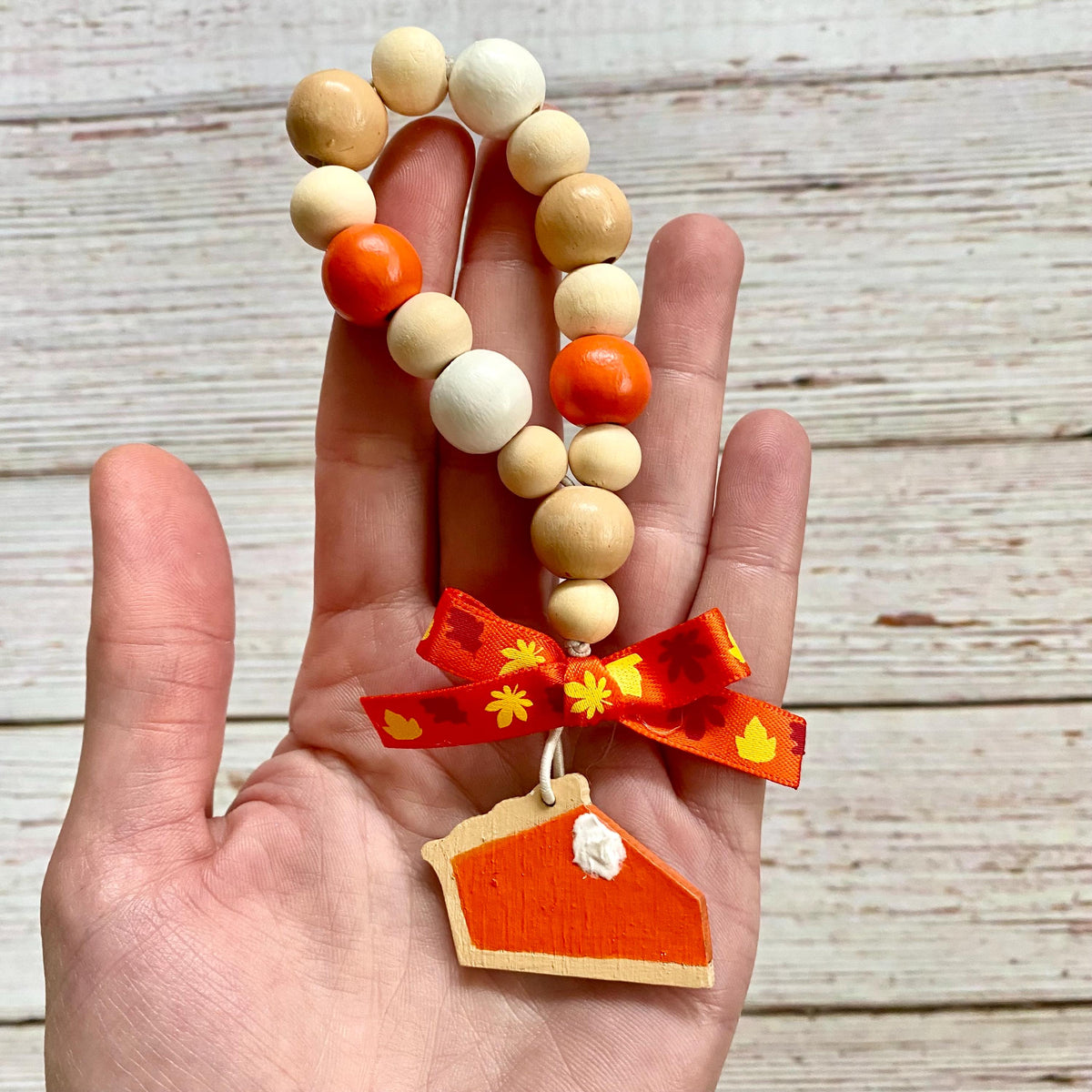 A hand holds a beaded loop made of natural wood beads with orange and white accents, tied with a red ribbon and ending in a pie slice charm, against a whitewashed wood background.