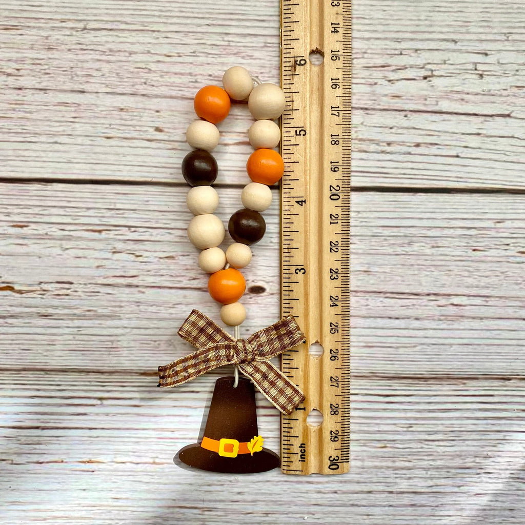 A decorative beaded garland with orange, cream, and brown wooden beads, a plaid bow, and a brown pilgrim hat ornament, shown next to a wooden ruler on a whitewashed wood background.