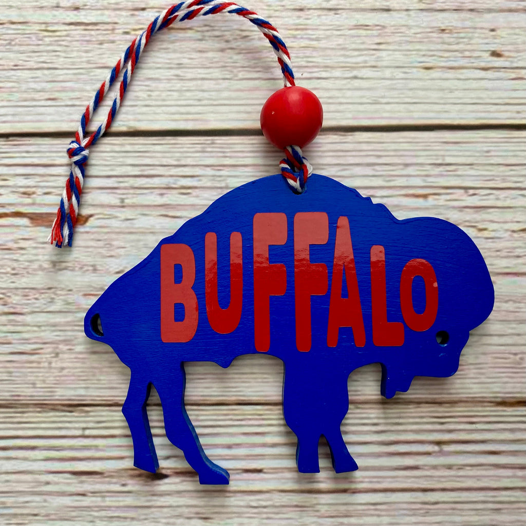 A blue buffalo silhouette hanging ornament with red 'BUFFALO' lettering, a red bead, and a red, white, and blue hanging string, shown on a light wood plank background.