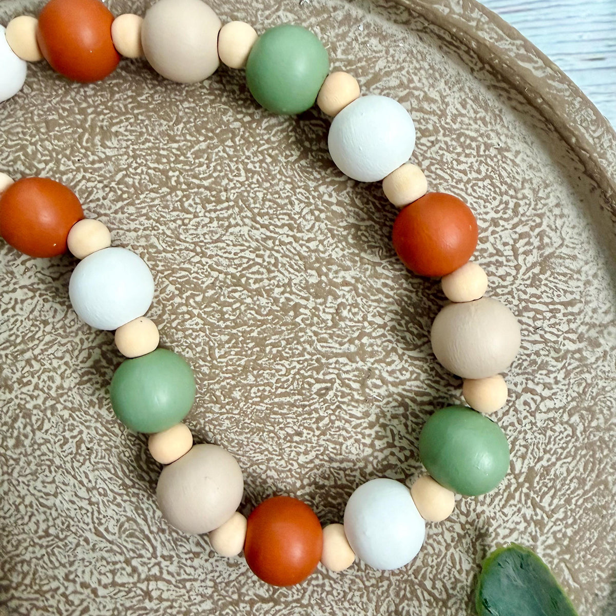 A beaded garland made of matte wooden beads in orange, sage green, cream, and white, arranged on a textured beige ceramic plate.