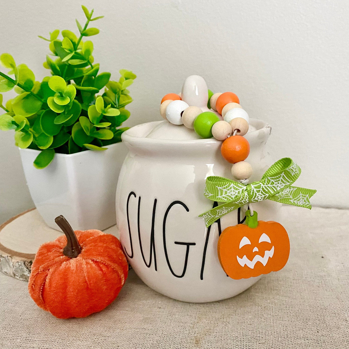 A white ceramic sugar bowl decorated with black lettering, a green and white beaded garland, a green bow, and an orange jack-o'-lantern ornament, styled on a beige fabric surface next to a small orange pumpkin and a potted green plant.