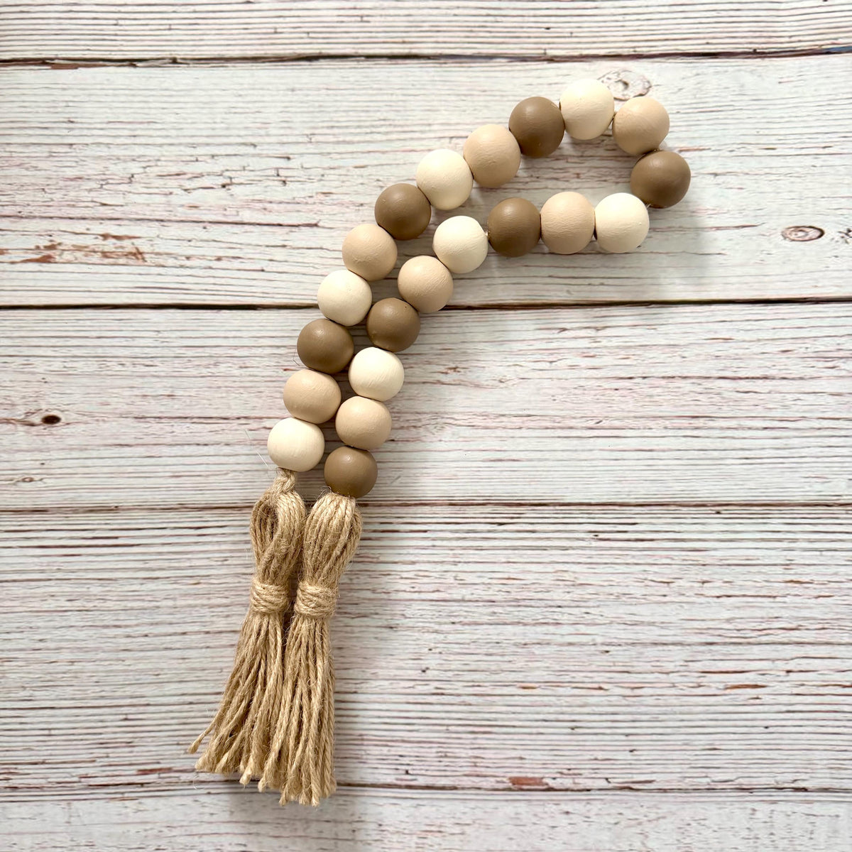 A wooden bead garland with alternating light beige, medium brown, and cream beads, finished with a natural jute tassel, laid flat on a rustic white-washed wooden background.