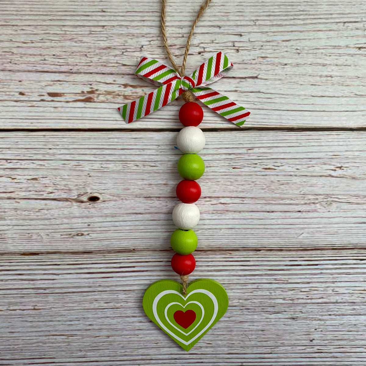 A hanging Christmas ornament made from a green wooden heart with layered white and red hearts, suspended below a red and green striped ribbon bow on a rustic white wood plank background.