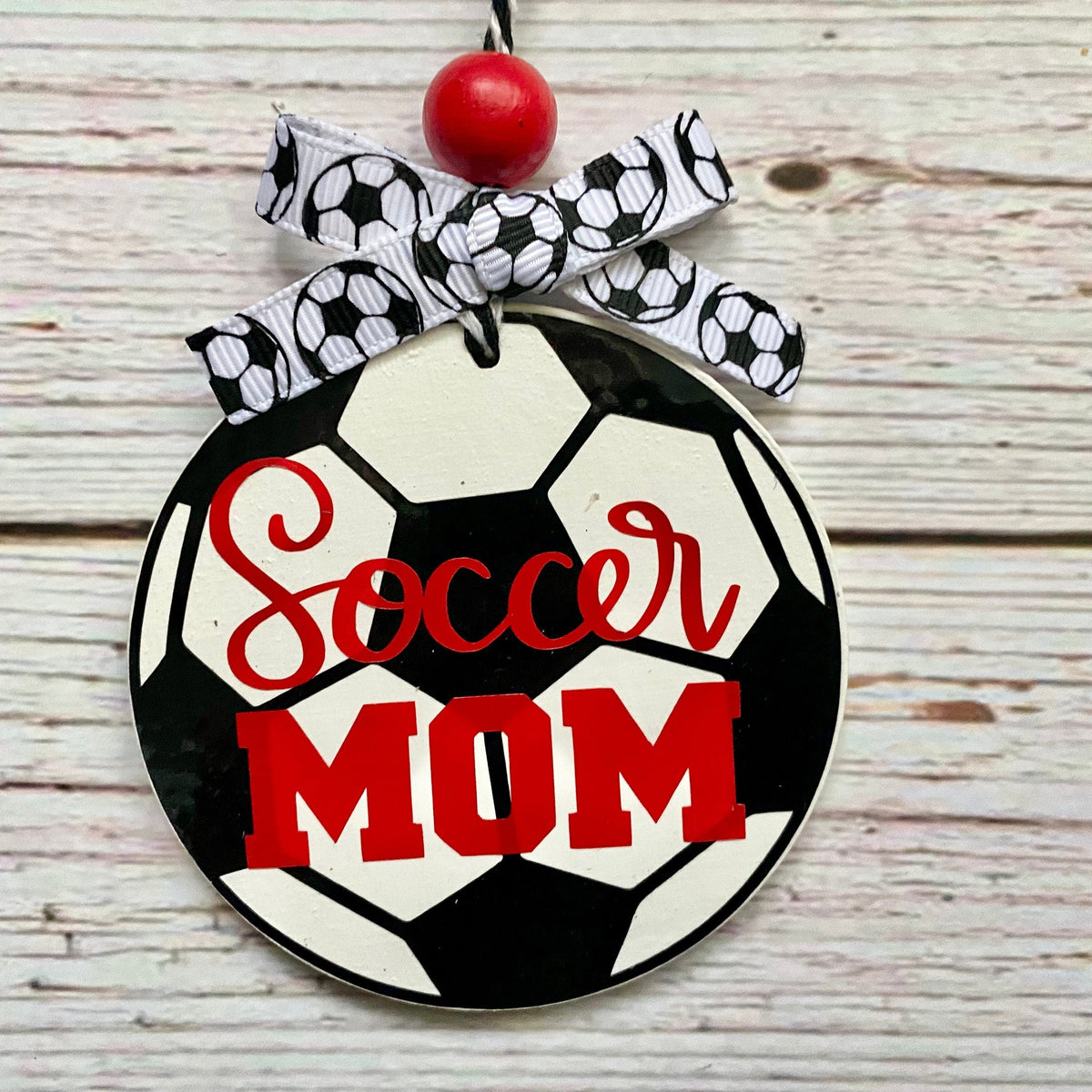 A round hanging ornament shaped like a soccer ball with the words 'Soccer MOM' in red script and block lettering, tied with a black-and-white soccer-pattern ribbon and a red bead at the top, shown on a whitewashed wood background.