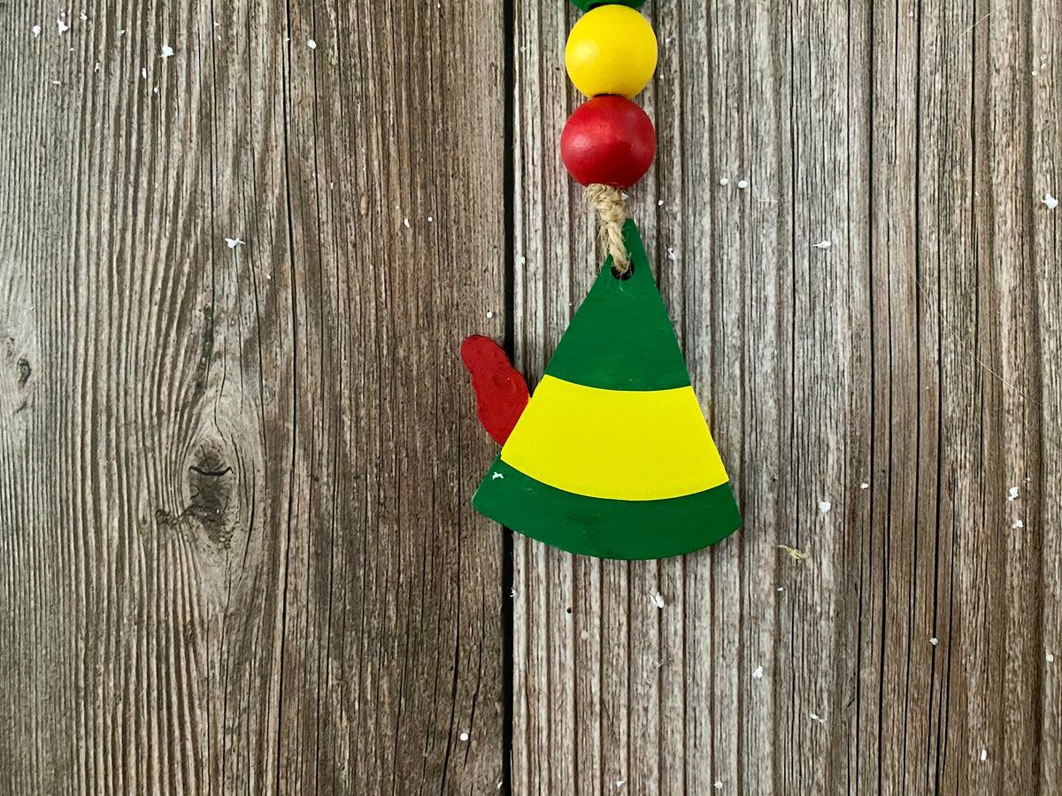 A handcrafted wooden Christmas elf ornament hanging on a rustic weathered wood background. The ornament features a green and yellow striped hat with a small red boot peeking out from the side, and a hanging string with red and yellow beads at the top.