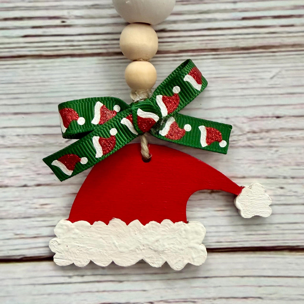 A Christmas Santa hat ornament made from a red wooden cutout with a white scalloped trim and a green ribbon bow printed with red and white Santa hats, hanging from wooden beads on a whitewashed wood background.