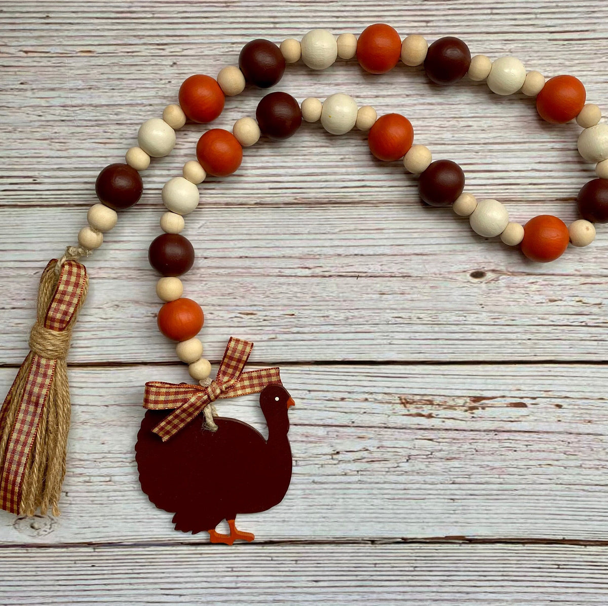 A rustic autumn-themed wooden bead garland featuring orange, dark brown, and cream beads, a matching gingham ribbon tassel, and a dark brown turkey silhouette ornament with a gingham bow, displayed on a whitewashed wood plank background.