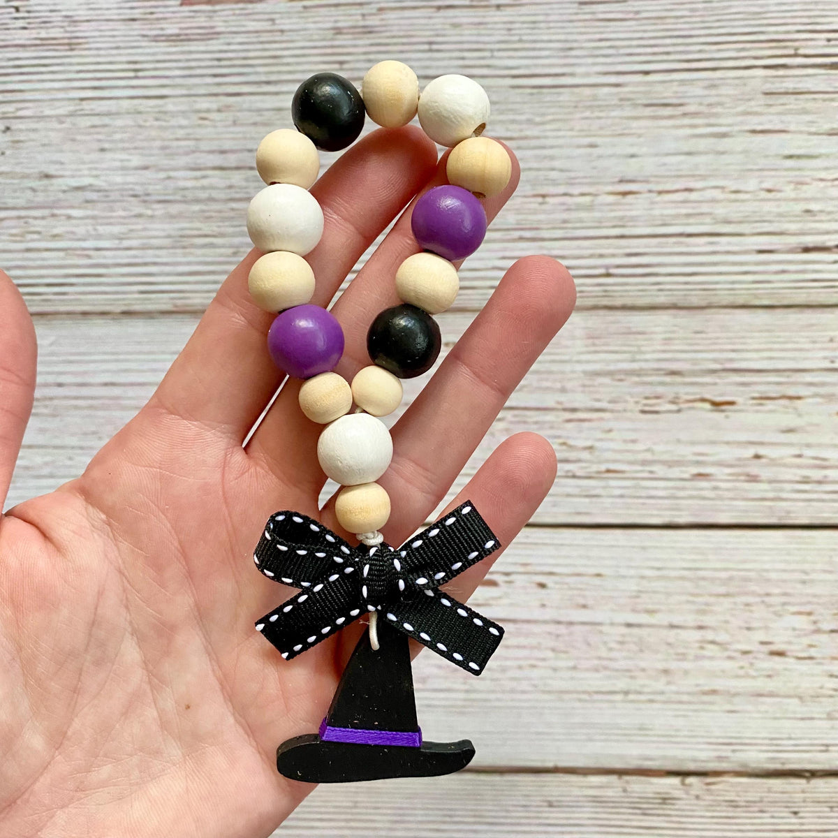 A hand holding a beaded garland with natural wood beads, purple and black beads, and a black witch hat charm with a black ribbon bow, set against a rustic whitewashed wood background.