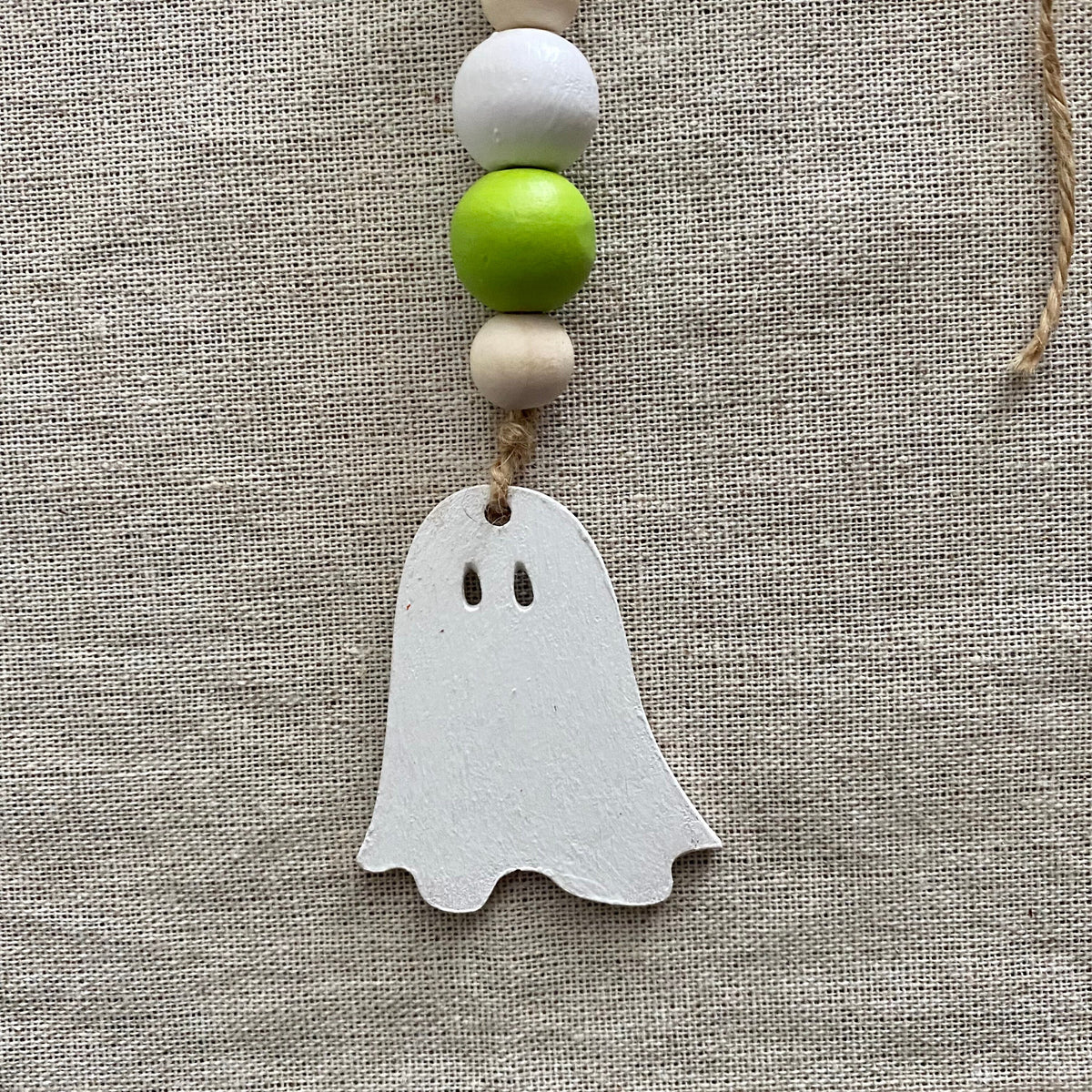 A white ghost-shaped wooden ornament hanging from a string with decorative beads, set against a textured beige linen background.