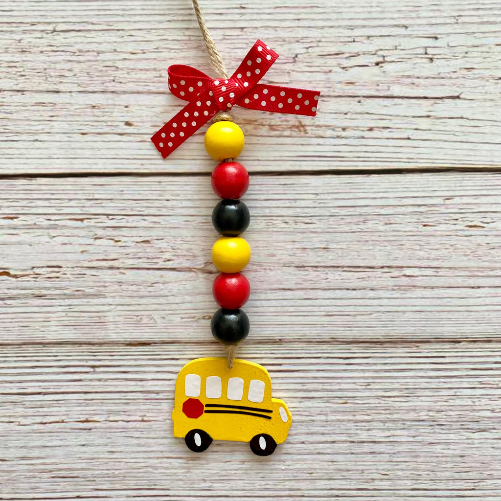 A decorative hanging ornament shaped like a yellow school bus, with a string of red, black, and yellow beads, a red polka-dot ribbon bow at the top, and a small red side mirror, shown against a whitewashed wood plank background.