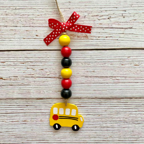 A decorative hanging ornament shaped like a yellow school bus, with a string of red, black, and yellow beads, a red polka-dot ribbon bow at the top, and a small red side mirror, shown against a whitewashed wood plank background.