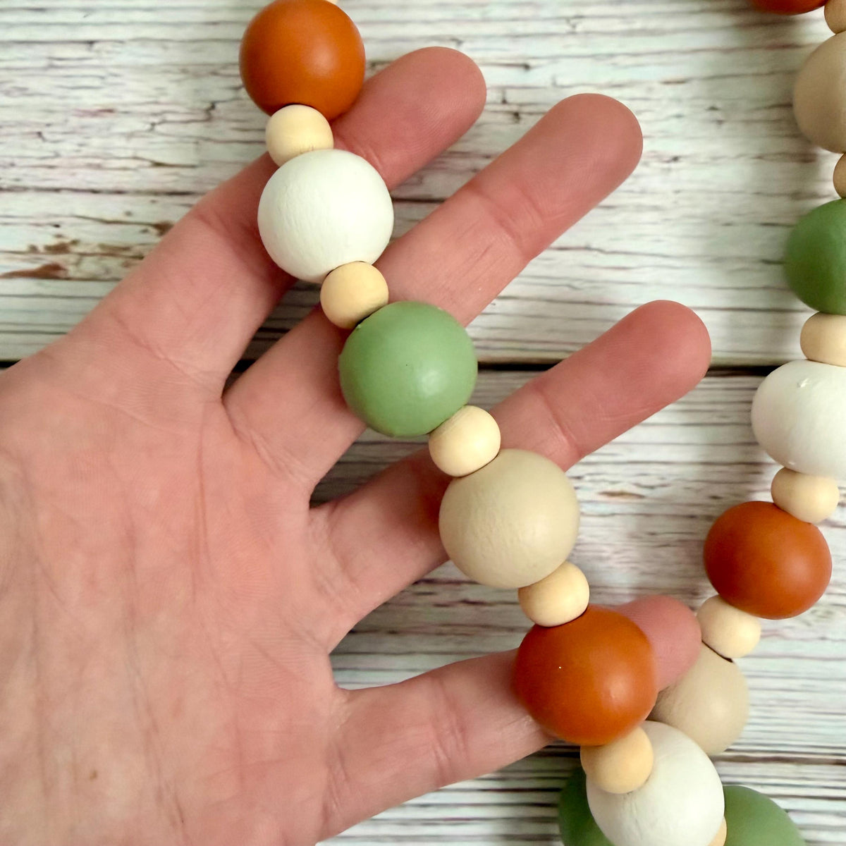 A close-up of a hand holding a wooden bead garland with matte orange, sage green, cream, and light tan round beads, set against a whitewashed wood background.