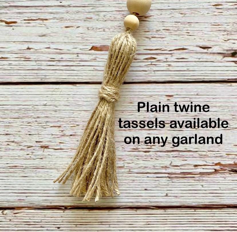 A close-up of a natural tan twine tassel with two small round wooden beads at the top, shown on a distressed white wood plank background with the text 'Plain twine tassels available on any garland'.