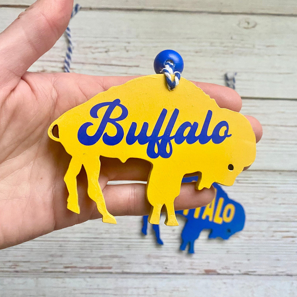 A hand holds a yellow buffalo-shaped ornament with blue 'Buffalo' lettering and a blue hanging loop, against a light wood plank background with a smaller blue ornament blurred behind it.