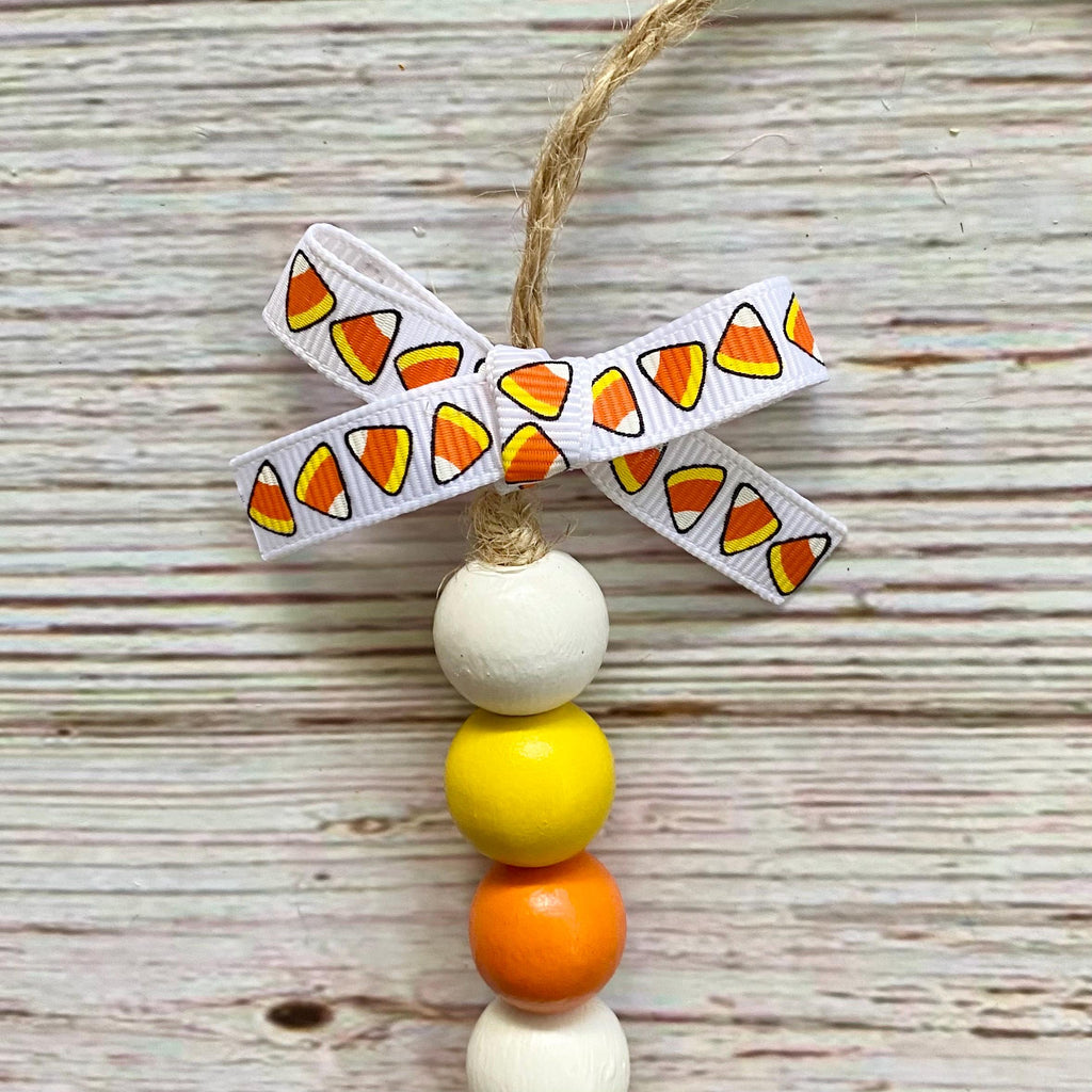 A handmade hanging ornament with a jute string, a white ribbon bow printed with candy corn motifs, and four wooden beads in white, yellow, orange, and white, displayed on a rustic whitewashed wood background.