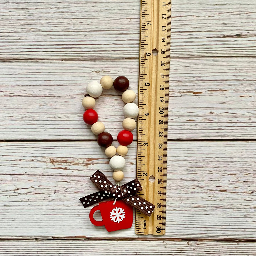 A wooden bead garland with red, cream, and dark brown beads, decorated with a brown polka dot bow and a red Christmas mug charm, shown next to a wooden ruler on a whitewashed wood background.
