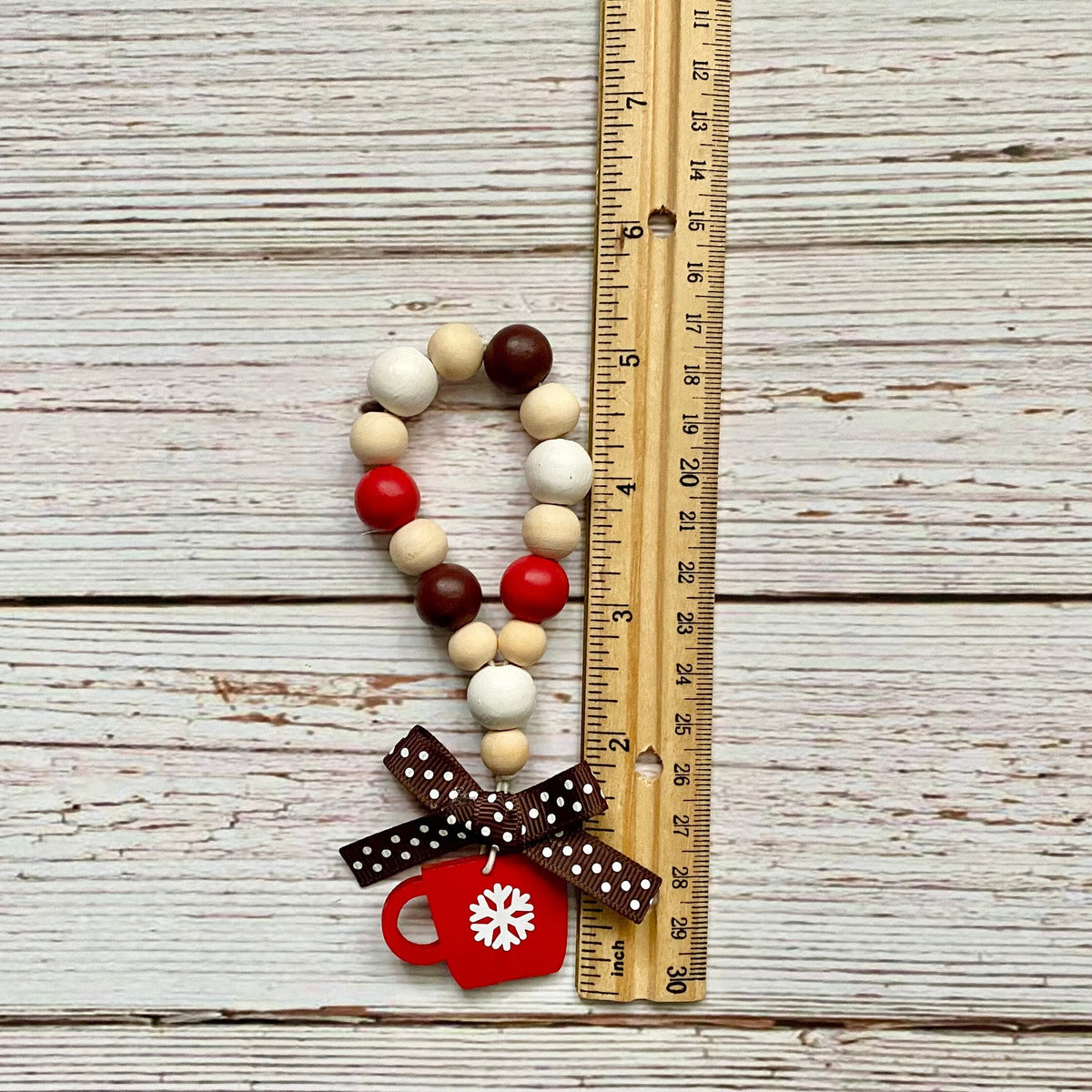 A wooden bead garland with red, cream, and dark brown beads, decorated with a brown polka dot bow and a red Christmas mug charm, shown next to a wooden ruler on a whitewashed wood background.