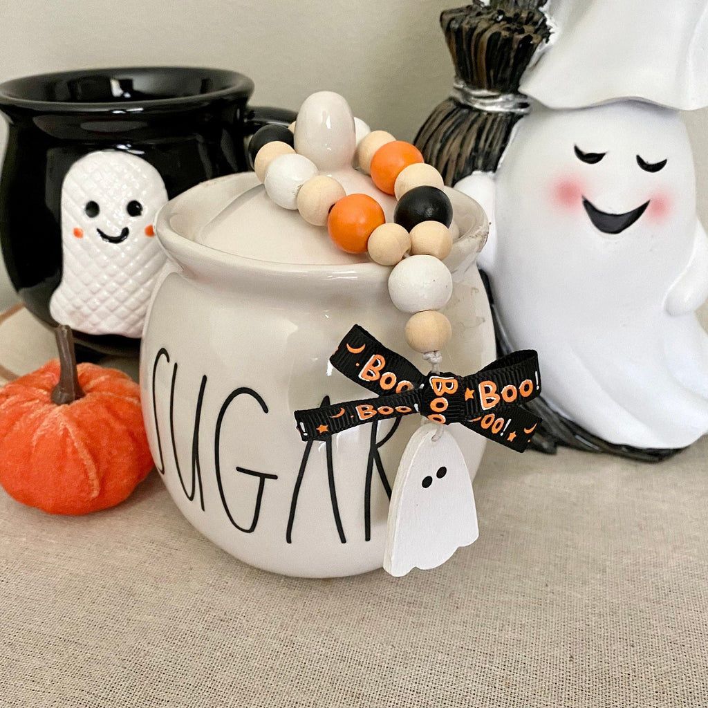 A white ceramic sugar jar decorated with a wooden bead garland, a black ribbon with 'Boo Boo 2021' text, and a ghost tag. It is styled on a beige fabric surface alongside a small orange pumpkin, a black cauldron, and a white ceramic ghost decoration.
