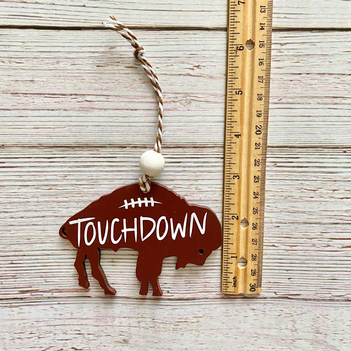 A brown buffalo-shaped metal ornament with the word 'TOUCHDOWN' in white, featuring a football graphic on its back, hanging from a twisted string with a white bead. The ornament is displayed next to a wooden ruler on a whitewashed wood background.