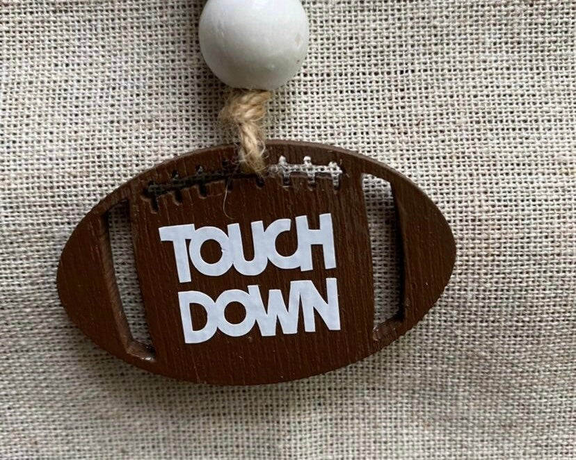 A wooden football-shaped ornament with 'TOUCH DOWN' painted in white letters, hanging from a rope with a white bead, set against a beige burlap background.