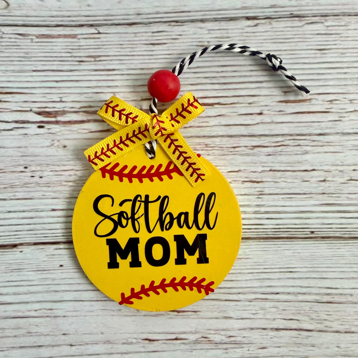 A round yellow softball-shaped ornament with 'Softball MOM' printed in black, decorated with red seam details, a yellow ribbon bow, and a red bead with a black-and-white string, shown on a whitewashed wood background.